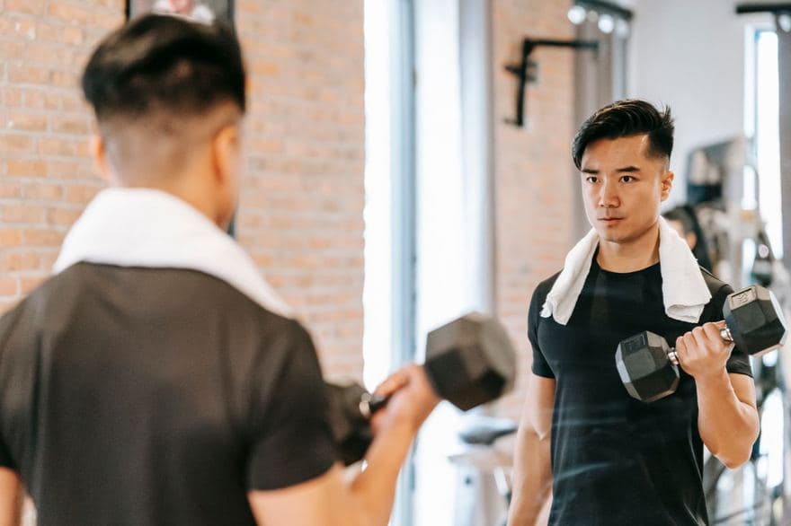 Một người đàn ông vắt khăn trên vai, cầm tạ tay tập trước gương trong phòng gym